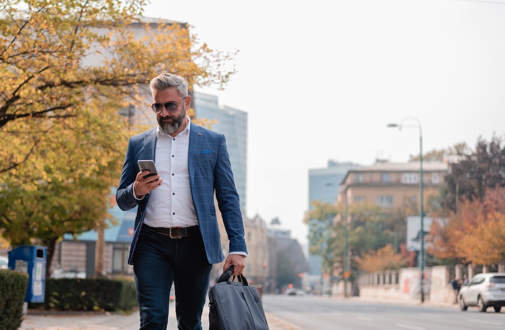 Businessman walking down a sidewalk while looking at his phone on a fall day.