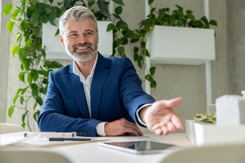 Man in a blue suit sitting at a desk and gesturing for someone to sit down.